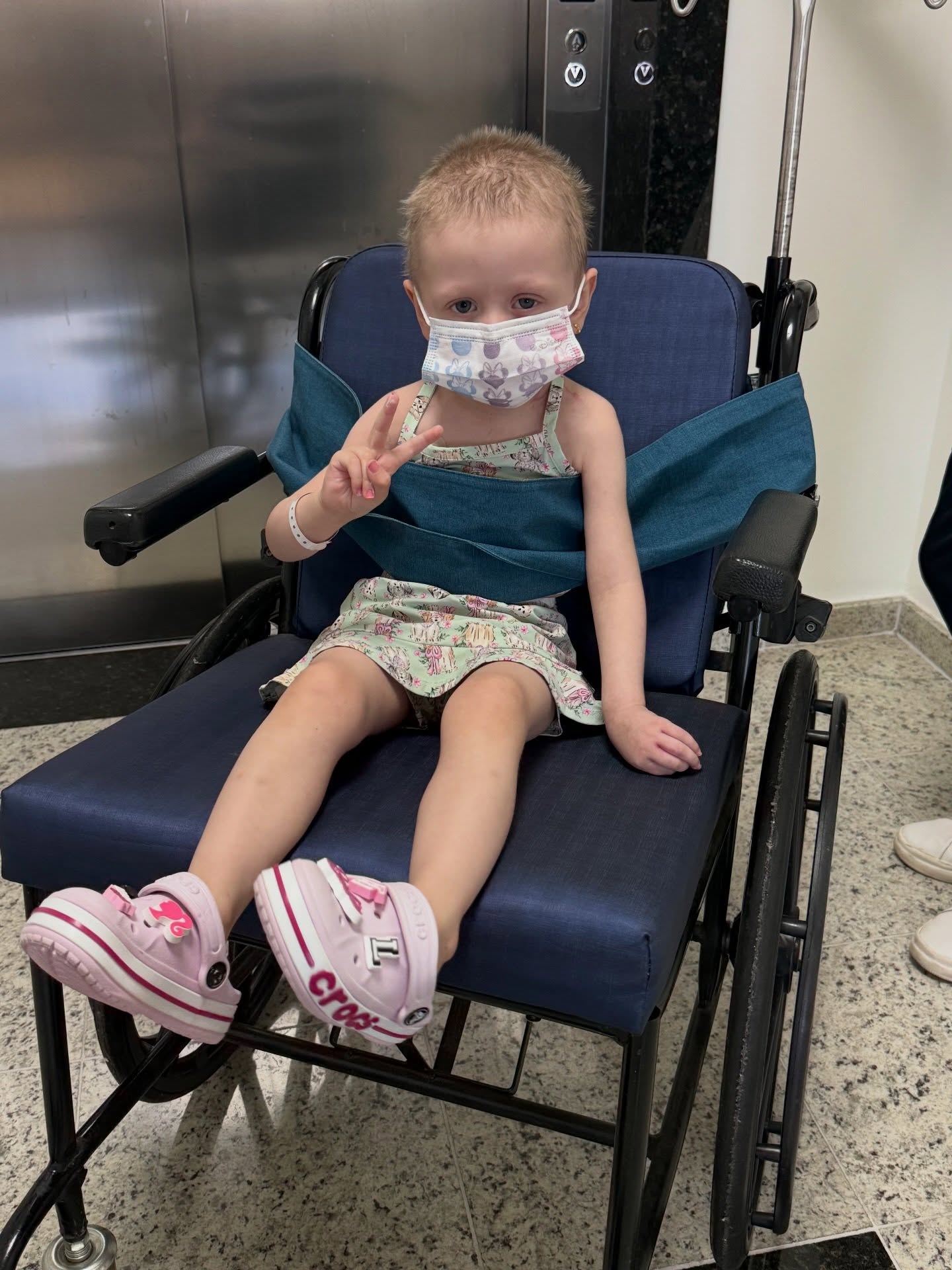 Olivia in a wheelchair wearing a face mask and a flowered dress, making a peace sign