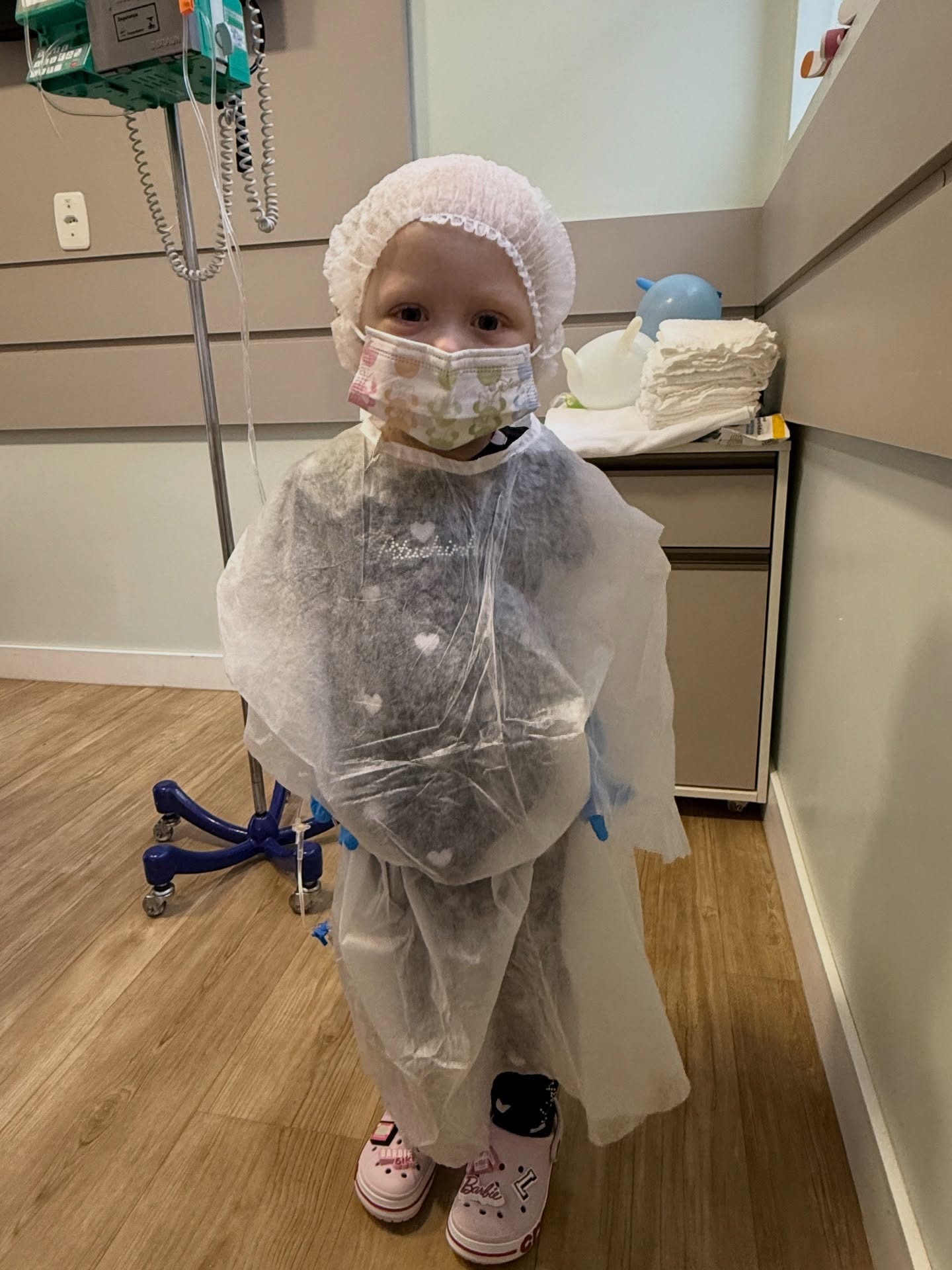 Olivia standing in a hospital room in a protective gown and cap, next to an IV pole